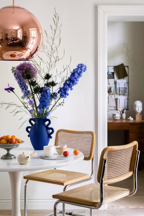 Unikatowe dekoracje w jadalni, prosty bialy stol, krzesla ratanowe i elegancka lampa nad stolem